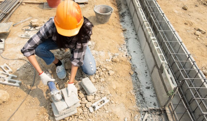 Trabalhadora com capacete e luvas utilizando ferramenta elétrica em canteiro de obras durante etapa de concretagem