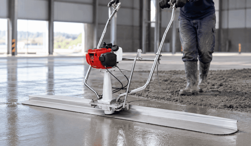 Operador em galpão industrial utilizando régua vibratória motorizada para adensamento preliminar e nivelamento de piso de concreto antes da locação de acabadora de superfície.